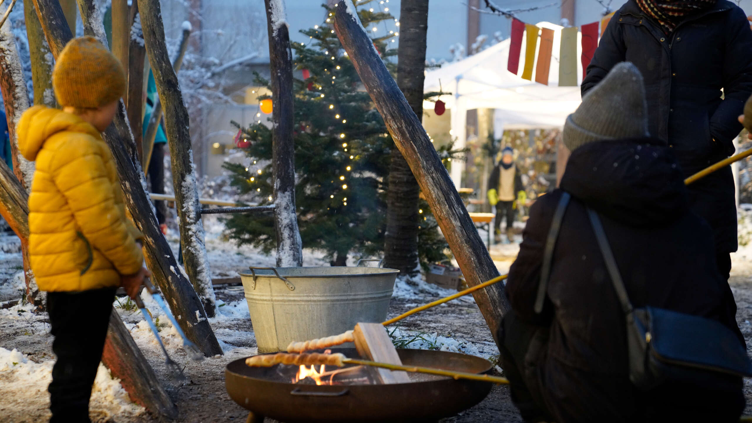 Weihnachtlich anmutender und winterlich verschneiter Pausenhof der Rudolf Steiner Schule Tannenbaum, Lichterkette und Menschen, die um eine Feuerschale herumstehen und Schlangenbrot am Feuer braten. Ein Junge steht daneben und im Hintergrund sieht man jemanden, der an einem Stand des Bazars arbeitet
