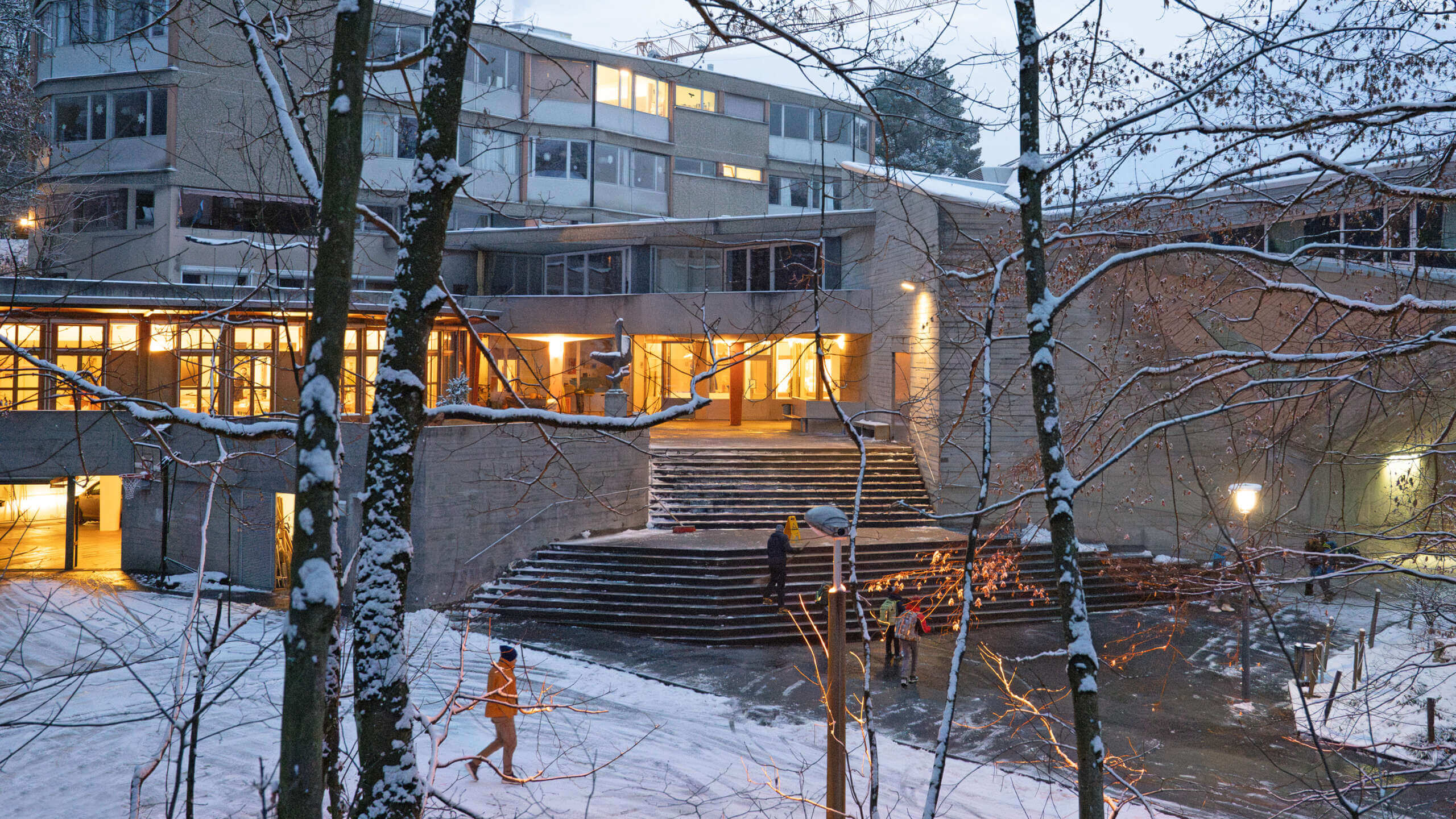 Winterlicher Vorplatz der Rudolf Steiner Schule Basel im Morgengrauen, mit Treppenaufgang, schneebedecktem Wendeplatz und warm beleuchtetem Schulhaus